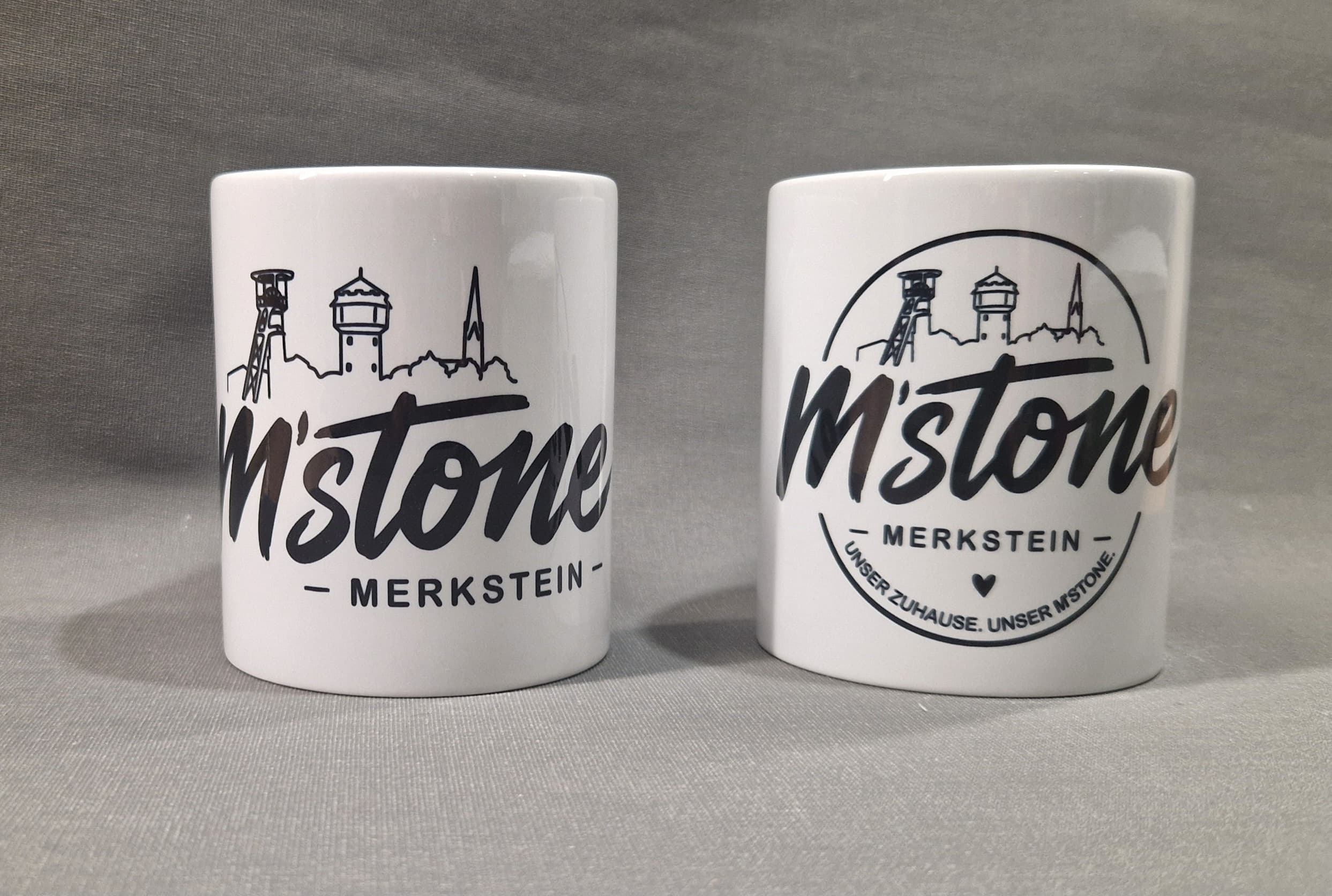 Zwei weiße M'stone Tassen: links mit Skyline-Schriftzug, rechts mit rundem Badge