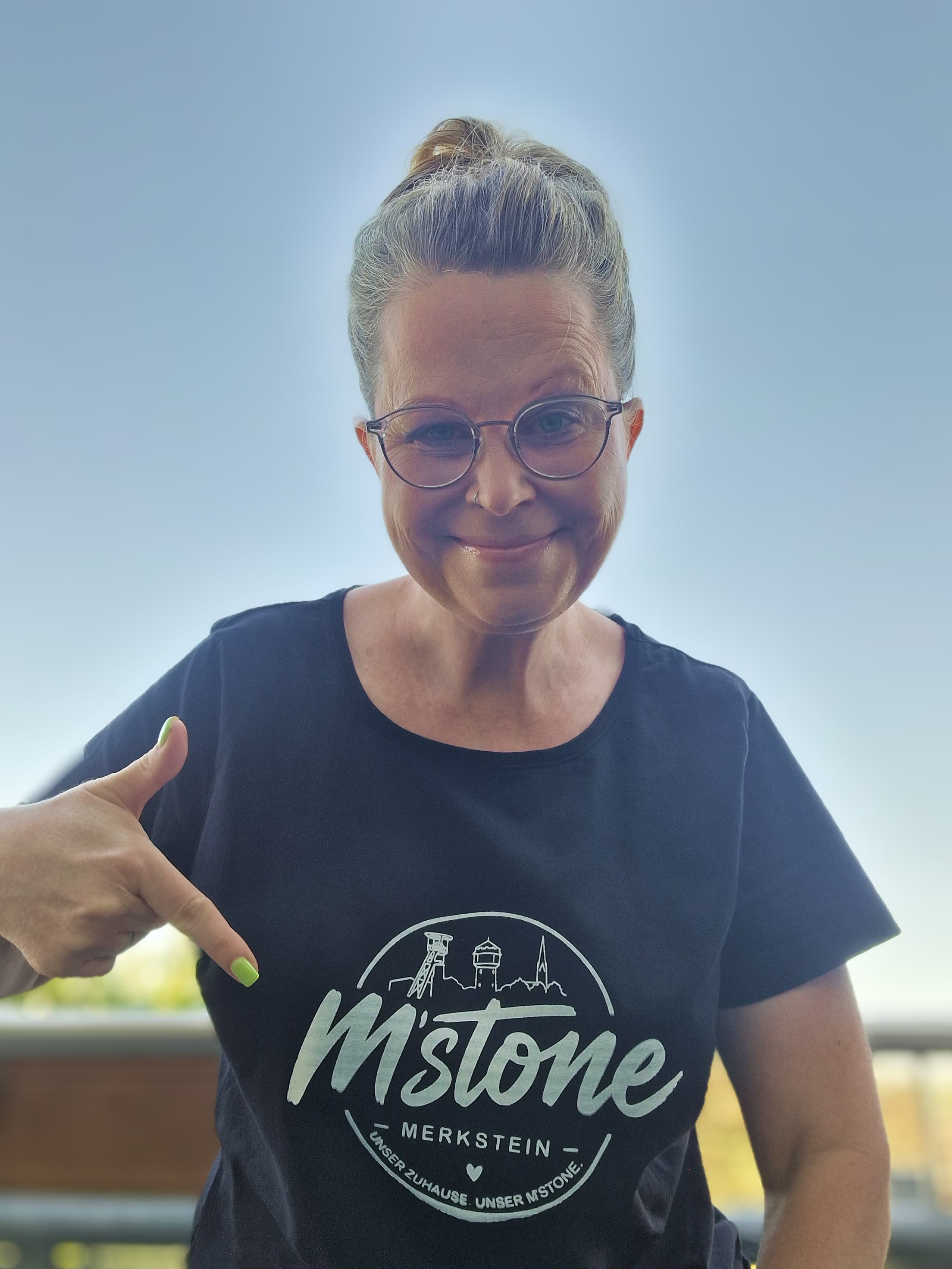 Merksteinerin im schwarzen M'stone T-Shirt zeigt stolz auf den Badge-Print